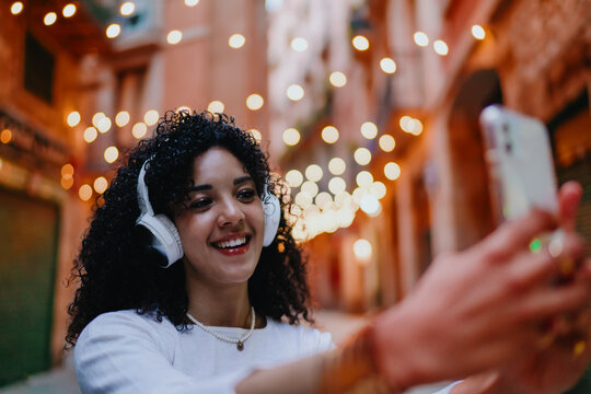Smiling young woman taking selfie photo on mobile phone and wearing wireless headphones while standing on cozy evening street with garland lights, digital technology lifestyle concept
