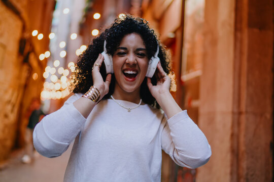 Happy young woman wearing white wireless headphones and laughing while standing on cozy evening street with garland lights, cheerful female student enjoying music podcast concept portrait
