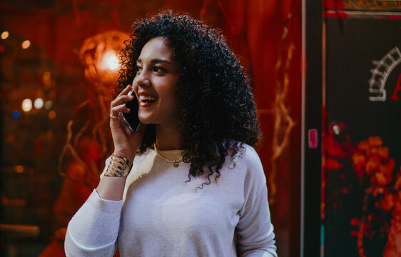 Smiling young woman talking on smartphone while standing near bright red illuminated restaurant showcase, happy female tourist enjoying mobile communication on evening city street