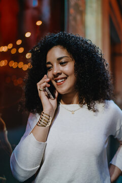 Cheerful young woman talking on smartphone and looking down while standing near bright cafe window on evening street, happy female tourist enjoying mobile communication portrait