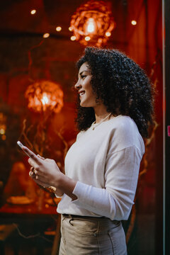 Smiling young woman holding smartphone and looking away near bright red illuminated restaurant showcase, cheerful female tourist enjoying evening city walk and mobile communication