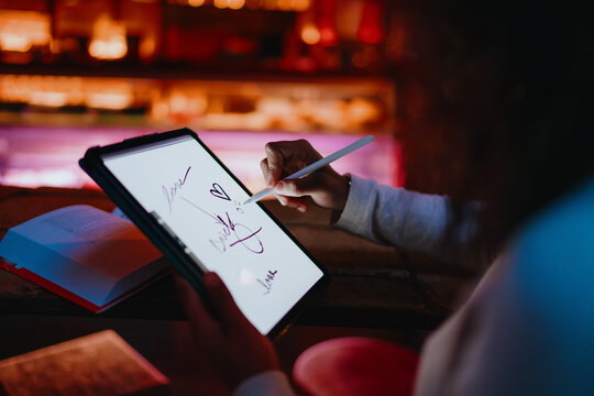 Close up of female hands holding smart stylus pen tool and writing notes on digital tablet screen inside cozy evening cafe, modern youth technology and remote learning concept