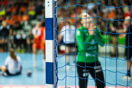 Handball goal net in sharp focus with blurred goalkeeper praing during professional indoor match, highlighting sports equipment and game atmosphere