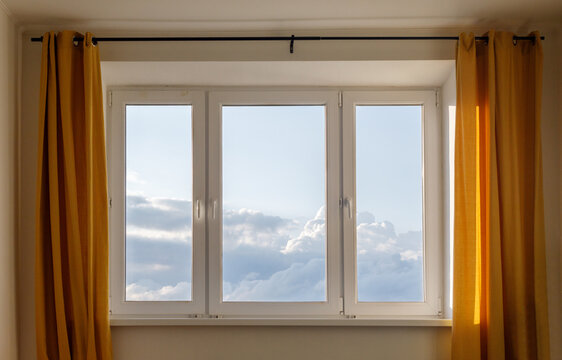 Interior view of a bright window with golden curtains overlooking a blue sky and clouds. Warm home interior and natural daylight creating a peaceful domestic atmosphere.