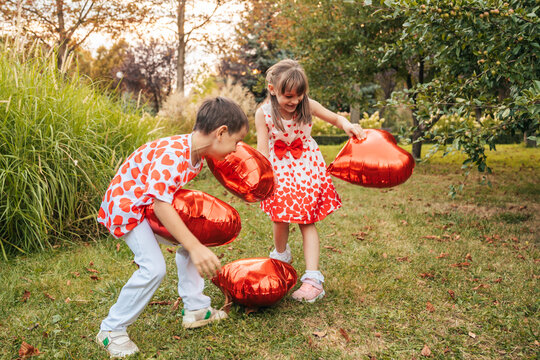 Two Children Play Outdoors With Red Heart-Shaped Balloons During Summer Break in a Garden Surrounded by Trees