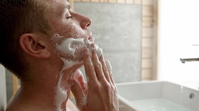 A young Caucasian man washing his face with cleansing foam in a bright modern home bathroom setting today.