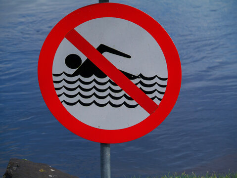No swimming sign stands near calm blue water. Symbol shows swimmer crossed out with red line. Sign warns against entering water for safety. Useful for beaches, pools, or lakes with restricted access