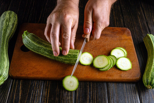 Hands skillfully slice a fresh zucchini on a wooden board, surrounded by more cucumbers. The warm kitchen provides a welcoming atmosphere for cooking