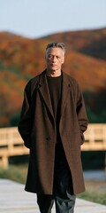 Obraz premium Mature man in a brown overcoat standing on a bridge with autumn hills portrait for fashion banner and editorial background