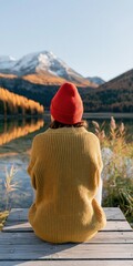 Fototapeta premium Person sitting on dock facing snow capped mountain over reflective lake in autumn with red beanie and mustard sweater for landscape wallpaper and travel poster