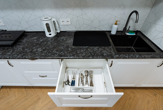 Top down view of modern kitchen countertop with open utensil drawer black sink and electric kettle near outlet, white cabinet interior and tiled backsplash in bright minimal home setting