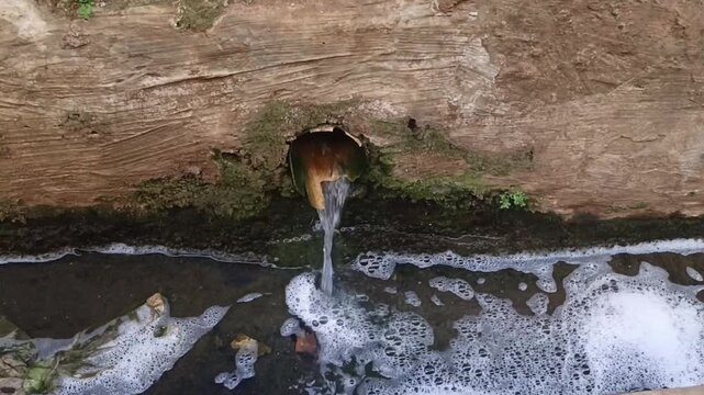 Water flows from a rustic metal pipe into a shallow drainage channel, creating foam and ripples against a mossy weathered wall.