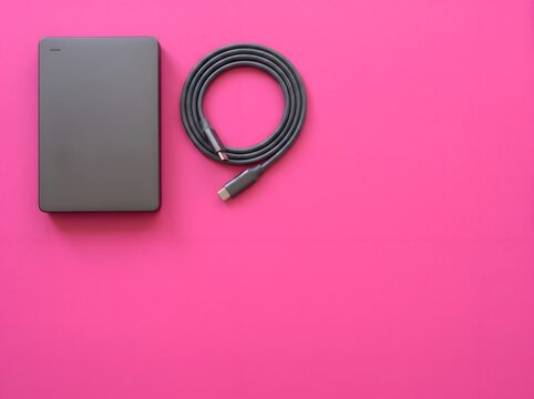A minimalist flat lay of a gray external hard drive and a coiled black USB cable on a vibrant pink background