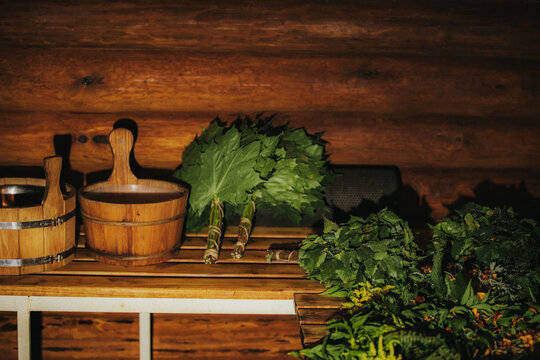 Wooden table displaying traditional sauna accessories including birch branches, wooden buckets, and various green leaves against a rustic log wall background