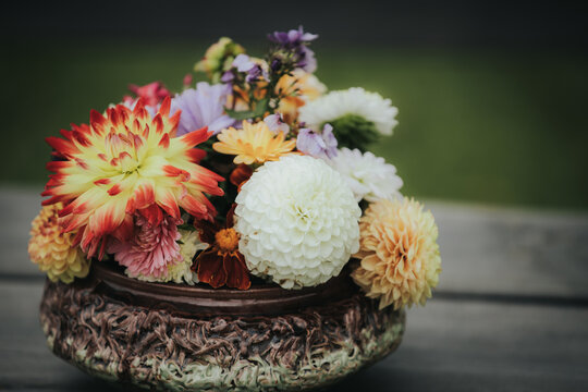 Colorful arrangement of various flowers including dahlias and zinnias in a decorative ceramic bowl placed on a wooden surface with green grass in the background