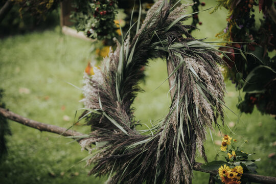 Decorative wreath made of dried grass and foliage hanging on a wooden structure in a garden setting with vibrant flowers and greenery in the background