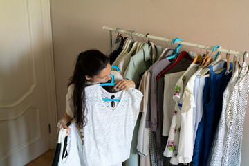 A woman looks closely at a sweater while holding it up next to other clothes in a room