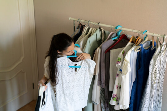 A woman looks closely at a sweater while holding it up next to other clothes in a room