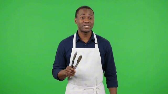 A man in an apron holding kitchen tongs, standing in front of a green screen background