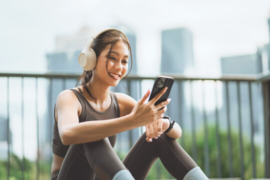 Smiling young woman in sportswear using smartphone and wearing headphones while resting after outdoor exercise, combining fitness.