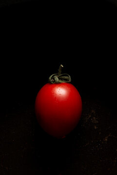 Single ripe red tomato with water droplets and a green stem placed on a black surface against a dark background.