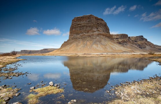 Large layered rock mountain reflected in a shallow pond beneath a blue sky with scattered clouds. lomagnupur mountain Iceland Landscape