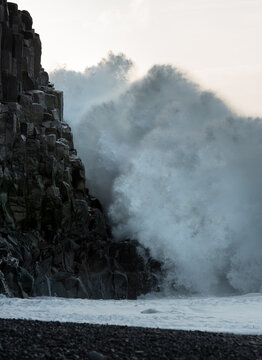 A massive ocean wave explodes in white sea foam against dark geometric basalt column cliffs on a black sand beach. Reynisfjara Iceland