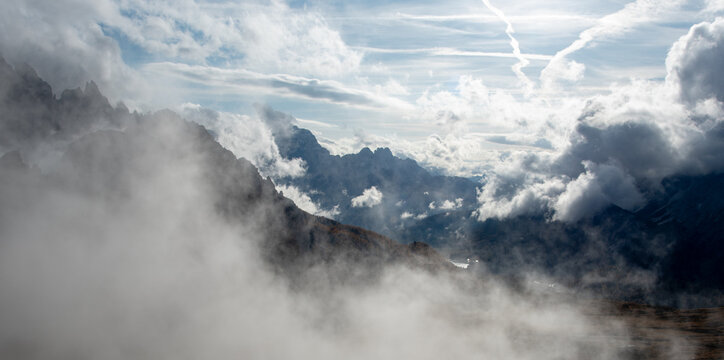 Jagged mountain peaks and a forested valley partly obscured by low clouds and mist beneath a bright sky. Tre Cime di lavaredo, Italy
