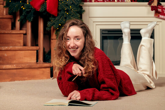 A cheerful young woman with curly hair relaxes on a carpeted floor near a decorated Christmas tree and fireplace, holding a pencil and open book