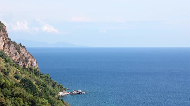 Rocky Green Coastline Overlooking Calm Blue Sea. Scenic rocky coastline with green vegetation overlooking calm blue sea and distant mountains in light haze.
