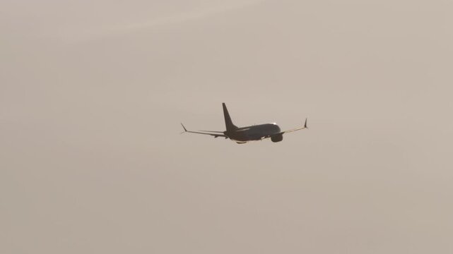 Commercial passenger airplane flies away into a hazy sky during golden hour sunset, telephoto shot of twin-engine jetliner ascending in mid-air