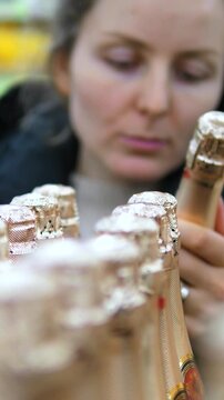 Woman selects champagne bottle from display. She leans in close, compares bottle tops, and grips one neck while other bottles remain lined up in front, creating refined retail tasting atmosphere.