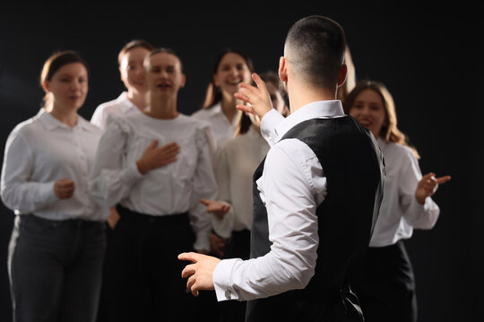 Choir leadership and musical performance, conductor leading a group of singers in a choral ensemble during a rehearsal on a dark stage background