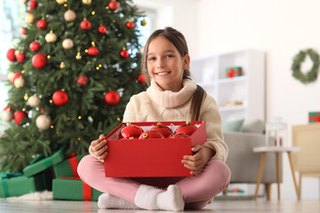 Smiling young girl holding a box of red christmas ornaments in front of a decorated christmas tree...
