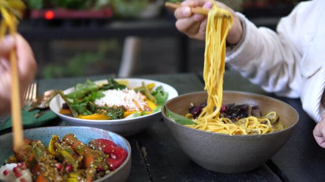 Person raises noodles with chopsticks from bowl, smiling in casual noodle meal setting. White sweater, dark table, and colorful shared dishes stay in view as noodles fall in long strands.