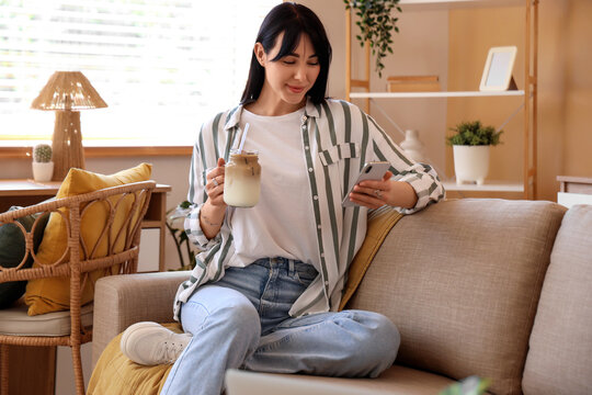 Young woman with glass jar of iced latte using mobile phone on sofa at home