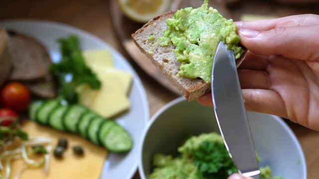 Hand spreads mashed avocado on bread, finishing open faced toast in homemade kitchen closeup. Precise knife work reveals relaxed breakfast prep with fresh vegetables and simple comfort food.