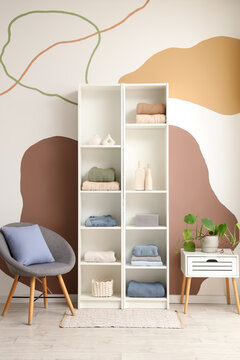 Shelving unit with clothes, stylish armchair and houseplant near color wall