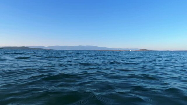 Low angle view of gentle sea waves rippling on deep blue water with distant islands and mountains under a clear sky in Ayvalik, Balikesir, Turkey.
