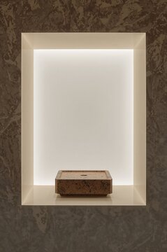 Small square brown stone sink basin centered inside a rectangular wall niche illuminated by soft hidden lighting against a dark marble wall