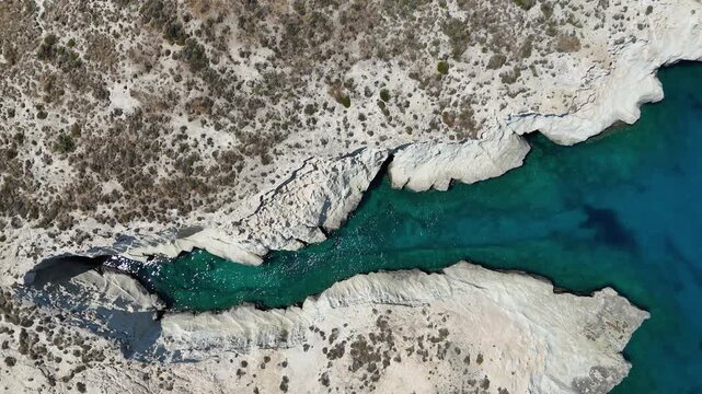 Aerial drone view looking down on the unique rocky landscape and the azure Aegean sea surrounding the Greek island of Milos in Greece. The ocean is clear blue. 