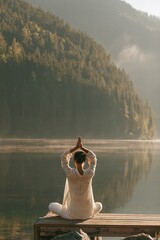 Fototapeta premium Woman doing yoga pose on a lakeside pier with mountain reflections for mindfulness background and wellness banner