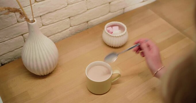 Female hand mixing warm chocolate drink with soft marshmallows on wooden table indoors