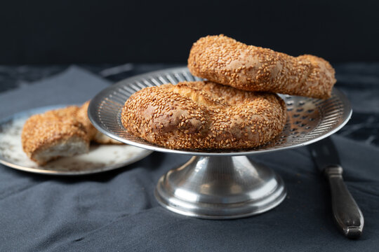 A close-up of freshly baked sesame rings arranged on a silver plate. The plate is on a table. The background is dark.
