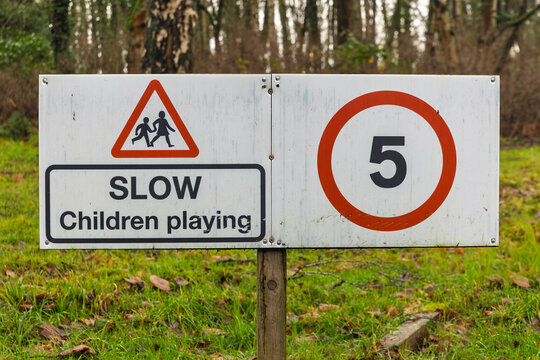 White metal sign combining red triangle hazard warning for children with slow children playing text panel, paired with circular 5mph maximum speed limit sign, mounted on wooden post in grassy woodland