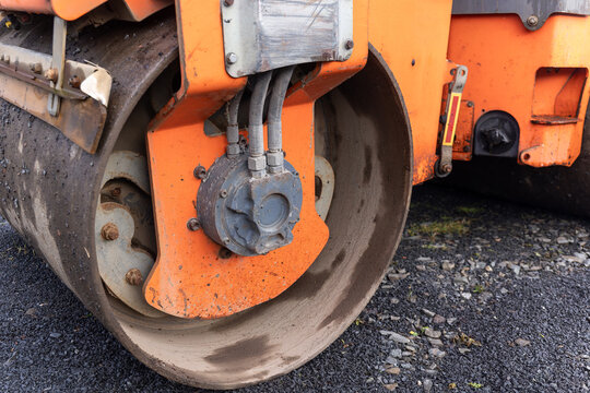 Heavy construction compaction machinery drum hub showing hydraulic motor, pressurised hoses and fittings mounted on orange steel frame, parked on freshly laid asphalt and gravel surface