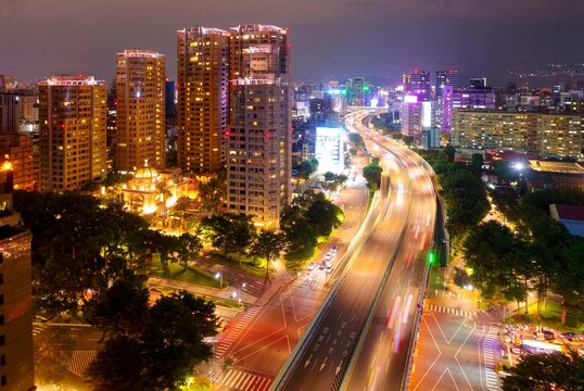 Aerial view of Jianguo S. Rd Expressway flying over Ren'ai Road, in Taipei, Taiwan, with residential towers of luxury apartments standing by the wooded avenue and city lights dazzling in the night