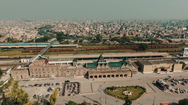Wide-angle drone perspective showcasing Rawalpindi Railway Station facade, green pitched roofs, and distant freight trains on parallel tracks in Pakistan.