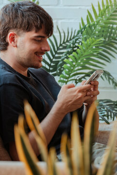 Smiling young man using a smartphone while relaxing at home surrounded by lush green houseplants, casual indoor lifestyle scene