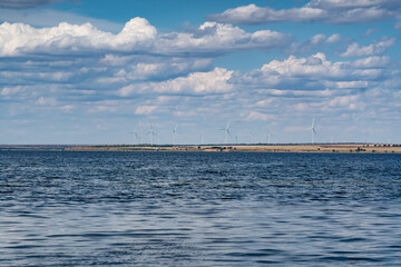 Fototapeta premium Wind turbines standing on the distant shore of a calm blue lake under a bright sky with fluffy white clouds, creating a peaceful countryside landscape.